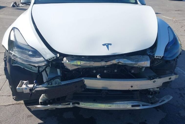 一輛白色特斯拉Model 3前端嚴重撞擊損壞，前保險桿完全脫落，大燈總成破損，引擎蓋變形，露出內部金屬結構框架。車事通提供專業的特斯拉Model 3事故車收購與評估服務，24小時全台到府服務。